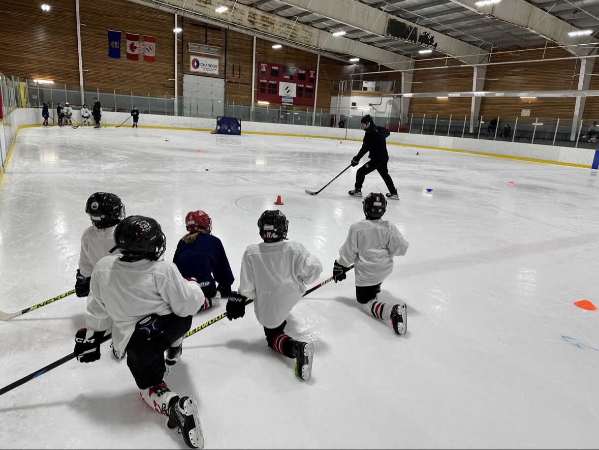 Young hockey players kneeling on the ice while a coach demonstrates a skill